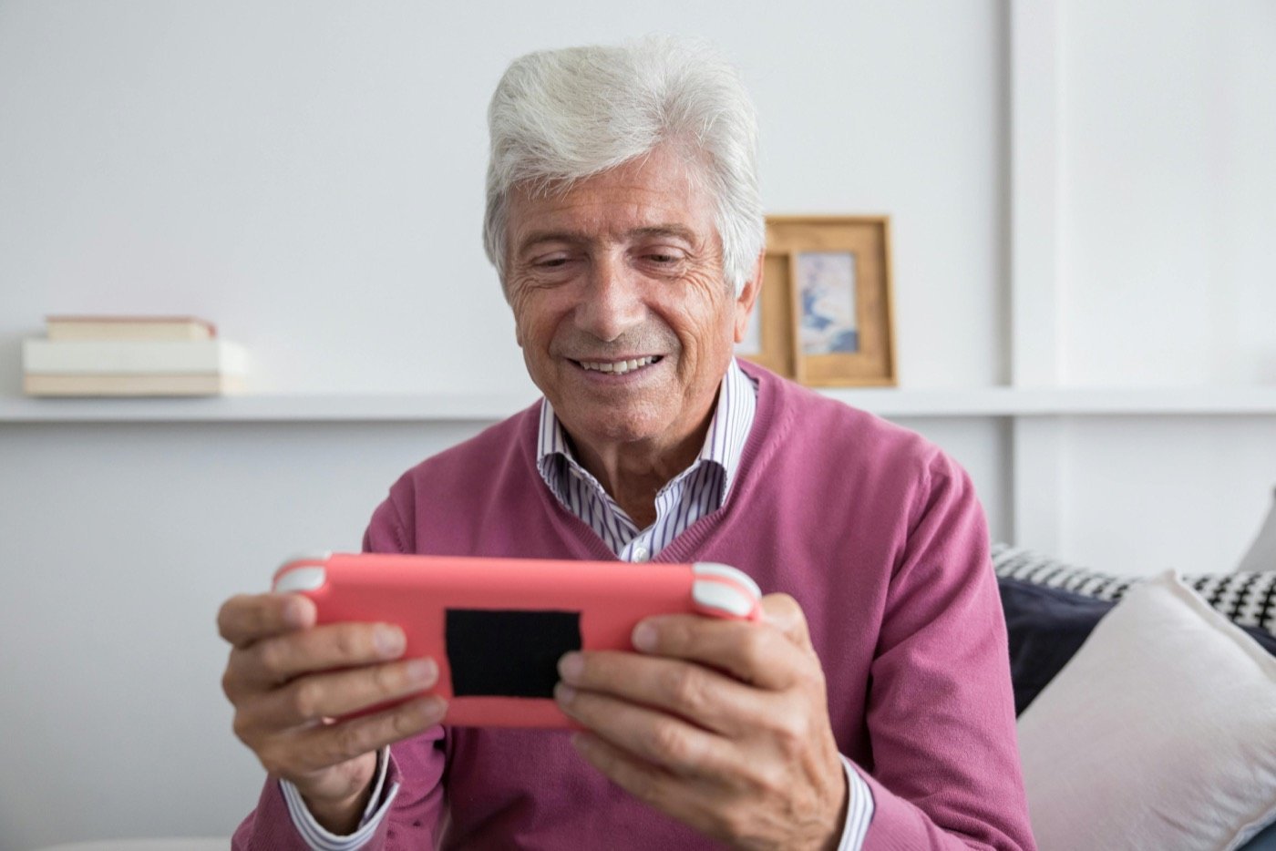 Un homme aux cheveux blancs sourit en tenant une console de jeu portable, assis dans son salon